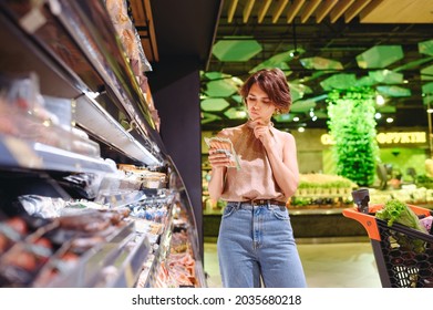 Young Minded Woman In Casual Clothes Shopping At Supermaket Greengrocery Store Buy Choose Meat Sausages Read Ingredients Shelf Life Prop Up Chin Grocery Cart Inside Hypermarke. Purchasing Food Concept