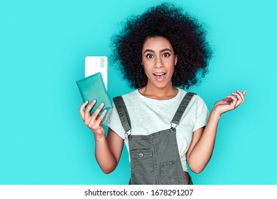 Young Mix Race Girl Wearing Denim Overall Standing Isolated On Blue Wall Holding Passport And Plane Ticket Looking Camera Surprised Unexpected Trip