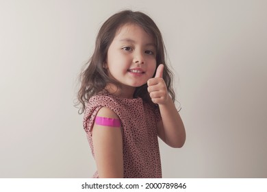 Young Mixed Asian Girl Giving Thumb Up And Showing Her Arm With Pink Bandage After Got Vaccinated Or Inoculation, Child Under 12 Years Old Immunization, Health Concept, Covid Delta Vaccine