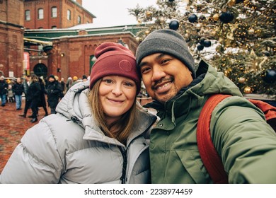 Young mixed couple taking a selfie wearing hat and jacket at a Holiday Christmas Market - Powered by Shutterstock