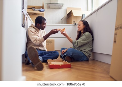 Young Mixed Ethnicity Couple Celebrating Moving Day In Kitchen Of New Home Eating Takeaway Pizza Sitting On Floor Surrounded By Removal Boxes Together