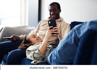 Young Mixed Ethnicity Couple Relaxing On Sofa At Home With Man Watching Tv Whilst Woman Lies Across Lap And Checks Social Media On Mobile Phone