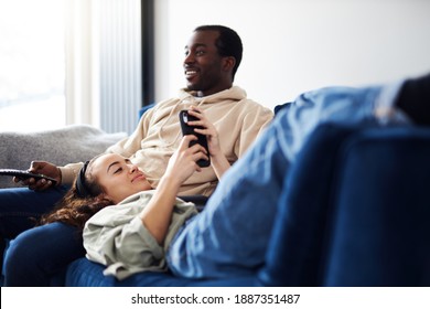 Young Mixed Ethnicity Couple Relaxing On Sofa At Home With Man Watching Tv Whilst Woman Lies Across Lap And Checks Social Media On Mobile Phone