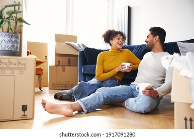 Young Mixed Ethnicity Couple Taking A Break On Moving Day Into New Home Sitting On Floor In Lounge Drinking Coffee Surrounded By Removal Boxes