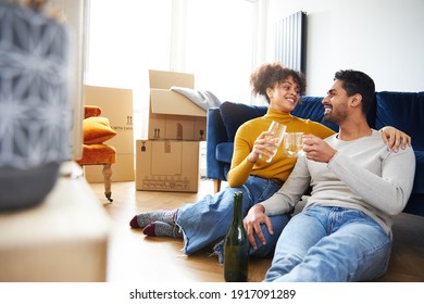 Young mixed ethnicity couple taking a break on moving day sitting on floor in lounge celebrating with champagne surrounded by removal boxes