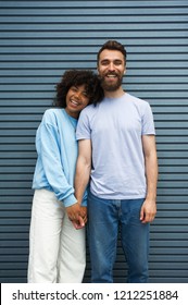 Young Mixed Hipster Couple In A Casual Clothes Posing Next To Each Other, Holding Hands, Smiling, Looking At Camera In An Urban Area On A Lovely Day. Afroamerican Girl