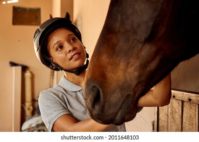 A young mixed race African woman blissfully smiling big and looking off camera while holding her horse’s face embracing it gently. She is wearing a grey shirt and black horse riding helmet - Powered by Shutterstock