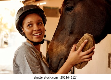 A Young Mixed Race African Woman Horse Owner Grooming Her Horse With A Hand Held Brush. She Is Standing In The Stables Wearing A Grey Shirt And Black Riding Helmet