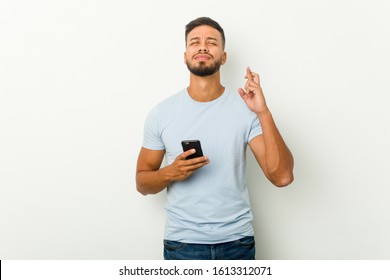 Young Mixed Race Asian Man Holding A Phone Crossing Fingers For Having Luck