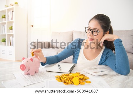 young mixed race asian woman deposit gold coin into piggy bank for future life sitting on sofa drinking coffee in the living room at home. interior and domestic housework concept.