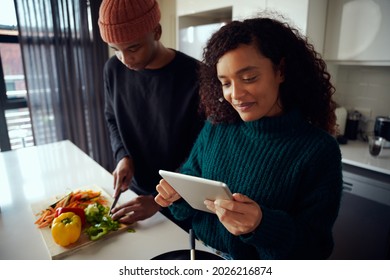 Young Mixed Race Couple Cooking Food In The Kitchen And Using A Tablet. Woman Looking At Tablet For Food Recipe. High Quality Photo