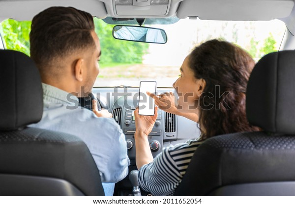 Young mixed race couple having car trip together,\
using mobile application on smartphone, shot from back seat,\
closeup. Brunette lady pointing at cellphone with empty screen,\
mockup