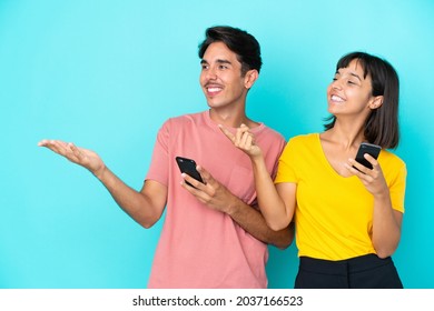 Young Mixed Race Couple Holding Mobile Phone Isolated On Blue Background Pointing Back And Presenting A Product