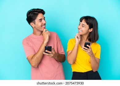 Young mixed race couple holding mobile phone isolated on blue background looking looking at each other