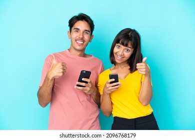 Young Mixed Race Couple Holding Mobile Phone Isolated On Blue Background Giving A Thumbs Up Gesture With Both Hands And Smiling