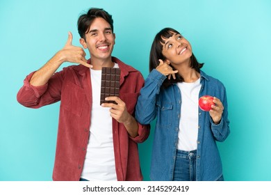 Young Mixed Race Couple Holding Apple And Chocolate Isolated On Blue Background Making Phone Gesture. Call Me Back Sign