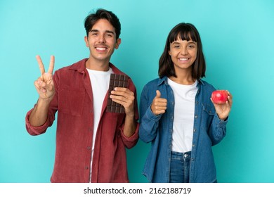 Young Mixed Race Couple Holding Apple And Chocolate Isolated On Blue Background Giving A Thumbs Up Gesture With Both Hands And Smiling