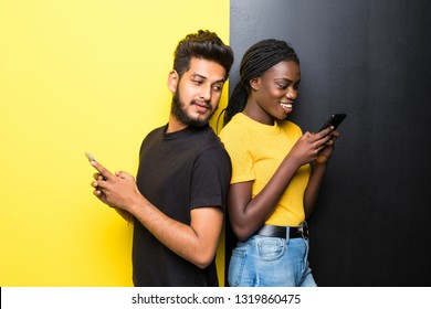 Young mixed race couple, indian man and african woman use phone standing back to back on the middle on different yellow and black background