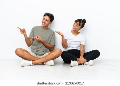 Young Mixed Race Couple Sitting On The Floor Isolated On White Background Pointing Back And Presenting A Product