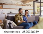 Young mixed race couple watching movie on laptop while relaxing on a weekend at home. Smiling indian man and beautiful woman sitting on sofa while using laptop and surfing the net.