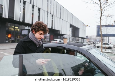 Young Mixed Race Curly Student Man In Black Coat With Smart Phone In Hands While Standing Near Small Car Over Shop Mall Outdoor In Cold Winter Weather.