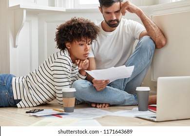 Young Mixed Race Family Couple Try To Solve Financial Situation, Study Business Papers, Plan Budget, Make Schemes On Laptop Computer, Drink Coffee, Pose On Floor In Living Room With Light Colours