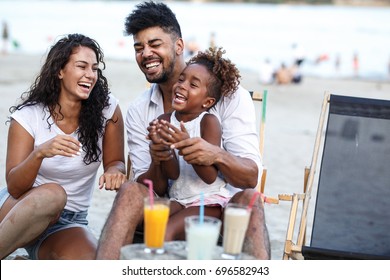 Young Mixed Race Family Sitting And Relaxing  At The Beach On Beautiful Summer Day.Daughter Lies In Father Lap And Laughing.