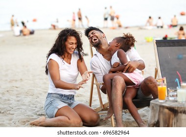 Young mixed race family sitting and relaxing  at the beach on beautiful summer day.Daughter lies in father lap and laughing.