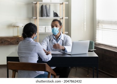 Young Mixed Race Male Doctor In Facemask Meeting With Female Patient, Listening To Complaints, Giving Consultation, Therapy Recommendation. Woman Visiting, Consulting Practitioner During Pandemic