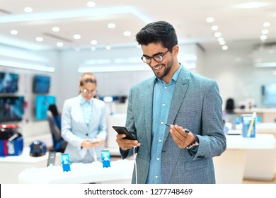 Young Mixed Race Man Dressed Smart Casual And With Eyeglasses Trying Out Smart Phone. Tech Store Interior.