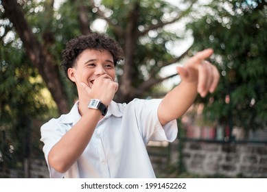 An young mixed race man points to someone, laughing out loud. Comedic situtation or mocking a person. Outdoor background.