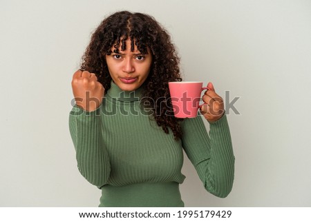 Young mixed race woman holding a mug isolated on white background showing fist to camera, aggressive facial expression.