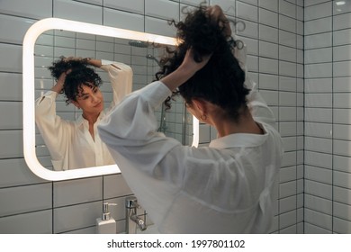 Young mixed race woman looking herself in the mirror in modern washroom. Girl makes a bun in her curly hair. Female making herself a ponytail. Morning routine concept. At home concept. - Shutterstock ID 1997801102