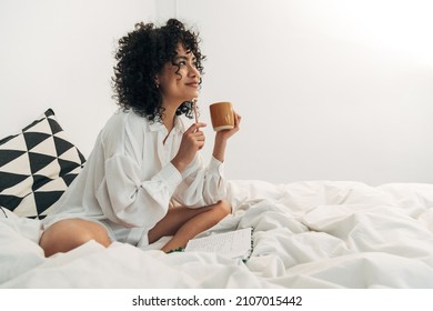 Young mixed race woman sitting on bed having coffee and writing on her journal. Copy space. - Powered by Shutterstock