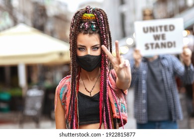Young mixed race woman wearing face mask showing peace sign during city street protest – Hipster female protesting outdoor for human rights while people holding unity sign in background