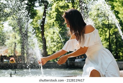 Young mixed race woman in white summer dress sitting by fountain on sunny day in park. Joyful positive woman cooling down by water on hot day outdoors.