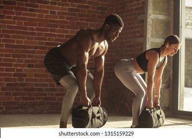 Young mixed-race fit couple doing squats exercise, while holding in hands heavy sandbag during group circuit functional training at the well-lit gym with panoramic windows.
