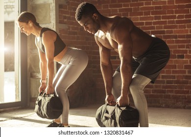 Young mixed-race fit couple doing squats exercise, while holding in hands heavy sandbag during group circuit functional training at the well-lit gym with panoramic windows. - Powered by Shutterstock