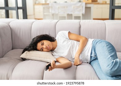 Young mixed-race girl in casual clothes resting lying down on the couch at home, feeling bored and sleepy, pointing with tv remote controller at camera, and switching channels, unhappy with program