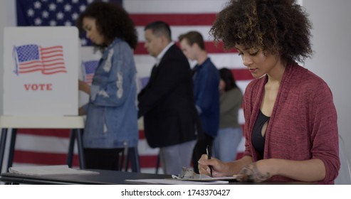 Young Mixed-race Woman Staffing Desk At Polling Station With Various Voters In Background, US Flag On Wall Behind Them.