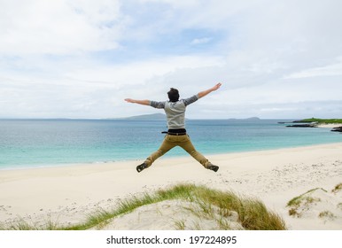 Young Model Jumping On A Sand Dune With Open Arms. White Sandy Beach And Blue Sky In The Background. Scotland, UK.