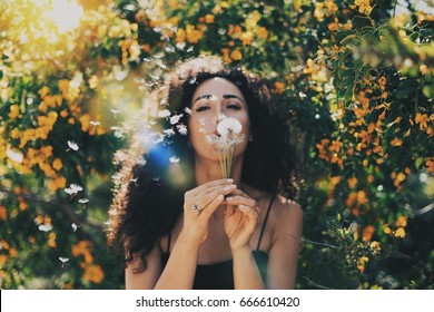 Young model look caucasian female with long dark curly hair wearing a black summer dress is blowing the dandelion at the camera while sitting on a blurred flowers background. Flare light. - Powered by Shutterstock