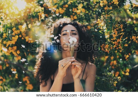 Young model look caucasian female with long dark curly hair wearing a black summer dress is blowing the dandelion at the camera while sitting on a blurred flowers background. Flare light.