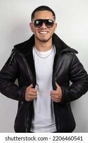 Young Model With Short Hair Wearing Sunglasses, Leather Sweater, White T-shirt And Necklace While Smiling, Urban Fashion And Style, Latin Man In Studio