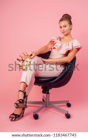 Young model woman in crown in a stylish summer outfit on pink background sitting on chair in studio