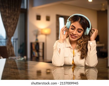 A Young modern attractive hipster happy Indian Asian woman female wearing trendy headphones is smiling and grooving while listening to music on an online streaming platform in an interior house setup.