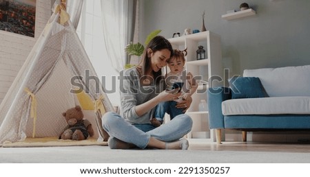 Young modern caucasian vlogger mom is showing her smartphone to little daughter, playing games with her or showing cartoons - family activity, technology concept 