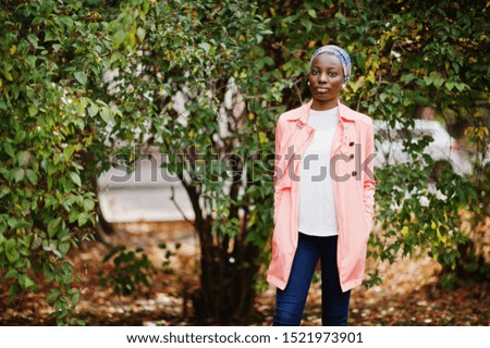 Young modern fashionable, attractive, tall and slim african muslim woman in hijab or turban head scarf and pink coat posed at park.