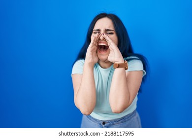 Young Modern Girl With Blue Hair Standing Over Blue Background Shouting Angry Out Loud With Hands Over Mouth 