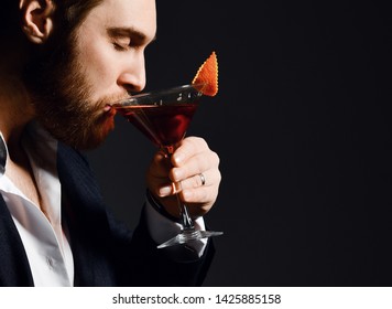 Young modern man bearded barman in fashion suit takes a sip of long awaited delicious red cocktail drink with orange in martini glass on dark background. - Powered by Shutterstock